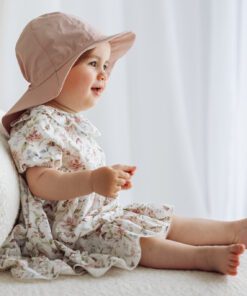 Baby girl wearing a floral dress and pink hat sits on a plush white sofa by a bright window.