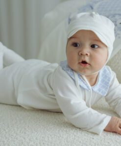 Baby in a white onesie and cap lying on a cream carpet propped on forearms and looking to the side