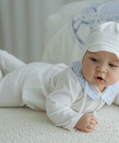 Baby in a white onesie and cap lying on a light colored rug and propping up on elbows looking to the side