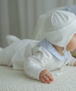 Baby in a white outfit and bonnet crawls on a pale textured bedsofa surface facing right