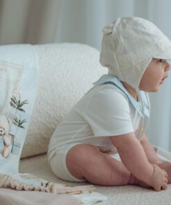 Baby wearing a light bonnet and white outfit sits on a cream sofa with a teddy bear blanket nearby