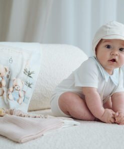 Baby in a white onesie and hat sits on a plush white couch beside a teddy bear themed blanket or towel with playful bears