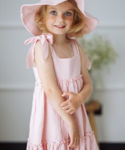 Little girl in a pink dress and wide brim hat smiling at the camera indoors