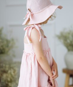 Young girl in a pale pink tiered sundress and matching wide brim hat with bows standing sideways indoors