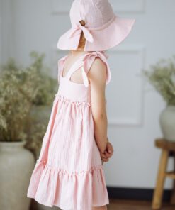 Young girl in a pink sleeveless dress with bow-tied straps and a large pink sun hat, standing sideways in a bright room.