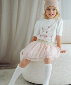 Smiling young girl in a pastel pink tutu skirt, white top, and beige knit hat sits on a white sofa, posing playfully.
