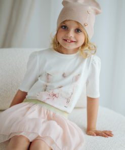 Smiling young girl in a beige knit hat, white top and pale pink skirt sits on a light sofa with a soft smile.