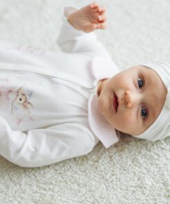 Baby girl lying on a light carpet in a white onesie with pastel embroidery and a white headwrap with a flower.