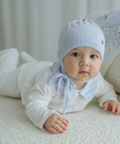 Baby lying on a soft cream carpet in a light blue knit hat and matching blue bib looking at the camera