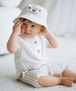 Cute baby sitting on a light carpet wearing a white hat with a bear face and cream striped shorts.