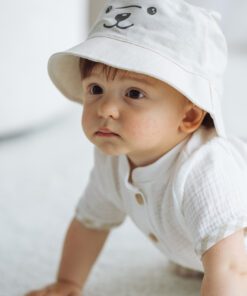 Baby crawling on a light carpet wearing a white hat with a bear-face design.