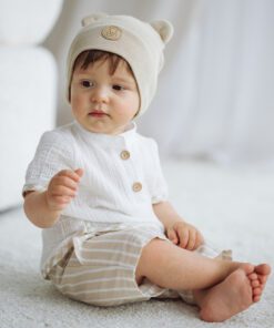Baby sitting on a light carpet wearing a cream knit hat with bear ears and a white top with beige striped shorts