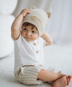Baby sitting on a light carpet wearing a cream bear ear hat and white top with beige striped shorts
