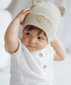 Baby wearing a cream knit hat with bear ears, looking to the side in a white outfit.