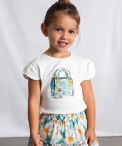 Portrait of a smiling young girl in a white shirt with a purse graphic and a teal floral skirt against a plain background