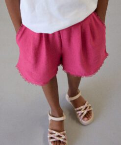 Child wearing pink shorts with scalloped hem white T shirt and beige sandals standing on a light floor