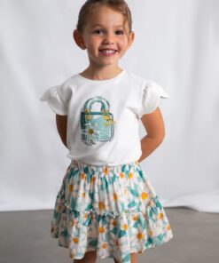 Smiling young girl in a white t shirt with a purse graphic and a teal daisy patterned skirt standing with hands on hips