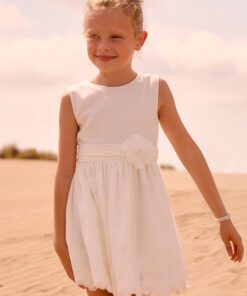 Young girl in a white dress standing on a sandy beach smiling at the camera under a sunny sky