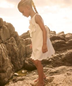 Young girl in a white dress with a flower sits on rocky shore at sunset looking down