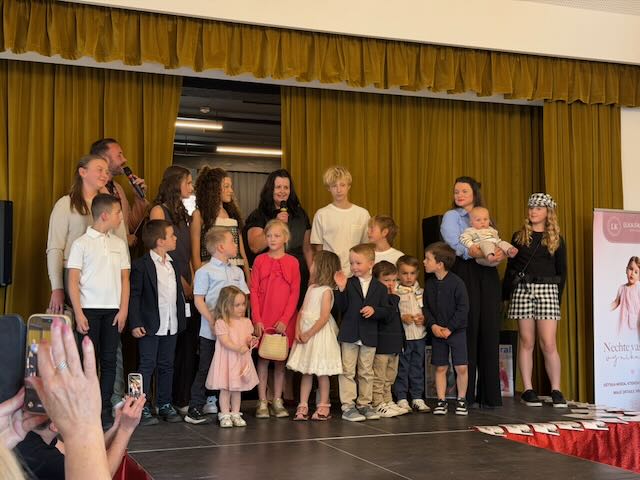Group of children and adults standing on a stage with yellow curtains posing for a photo at a ceremony or event